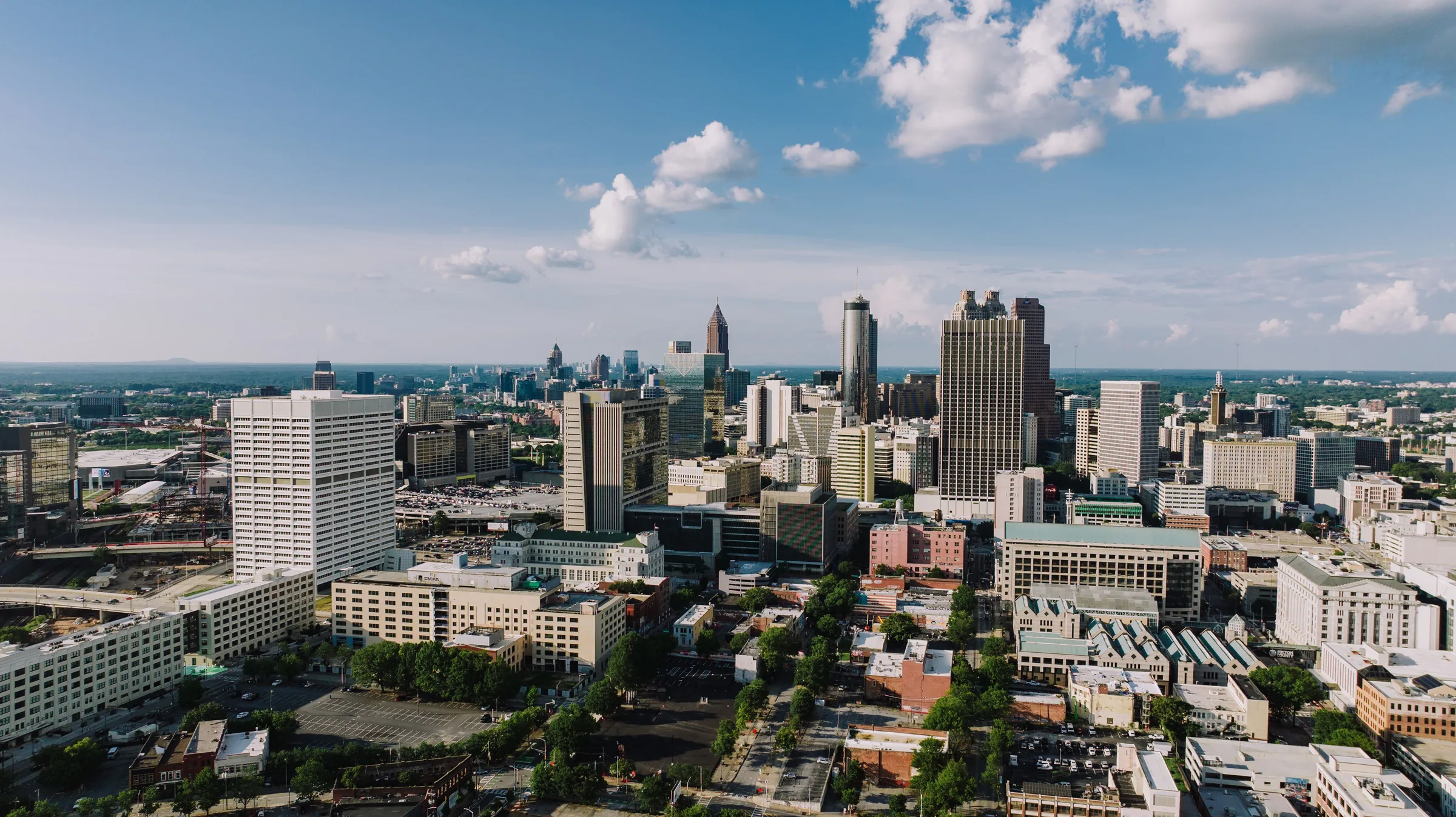 Aerial view of Atlanta skyline with downtown skyscrapers, green trees, and blue sky with white clouds.