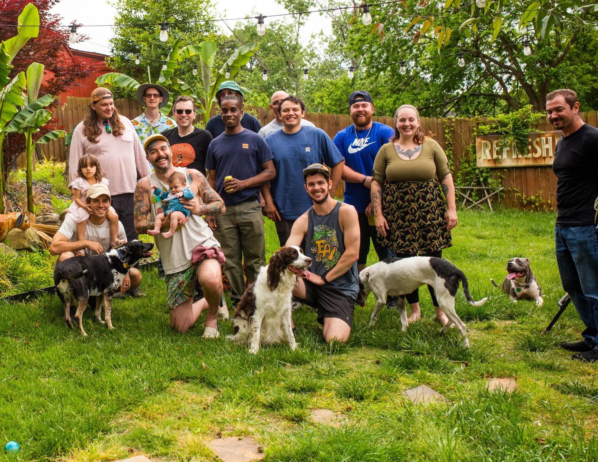 Large group of people and dogs gathered in a lush backyard with green lawn and trees.
