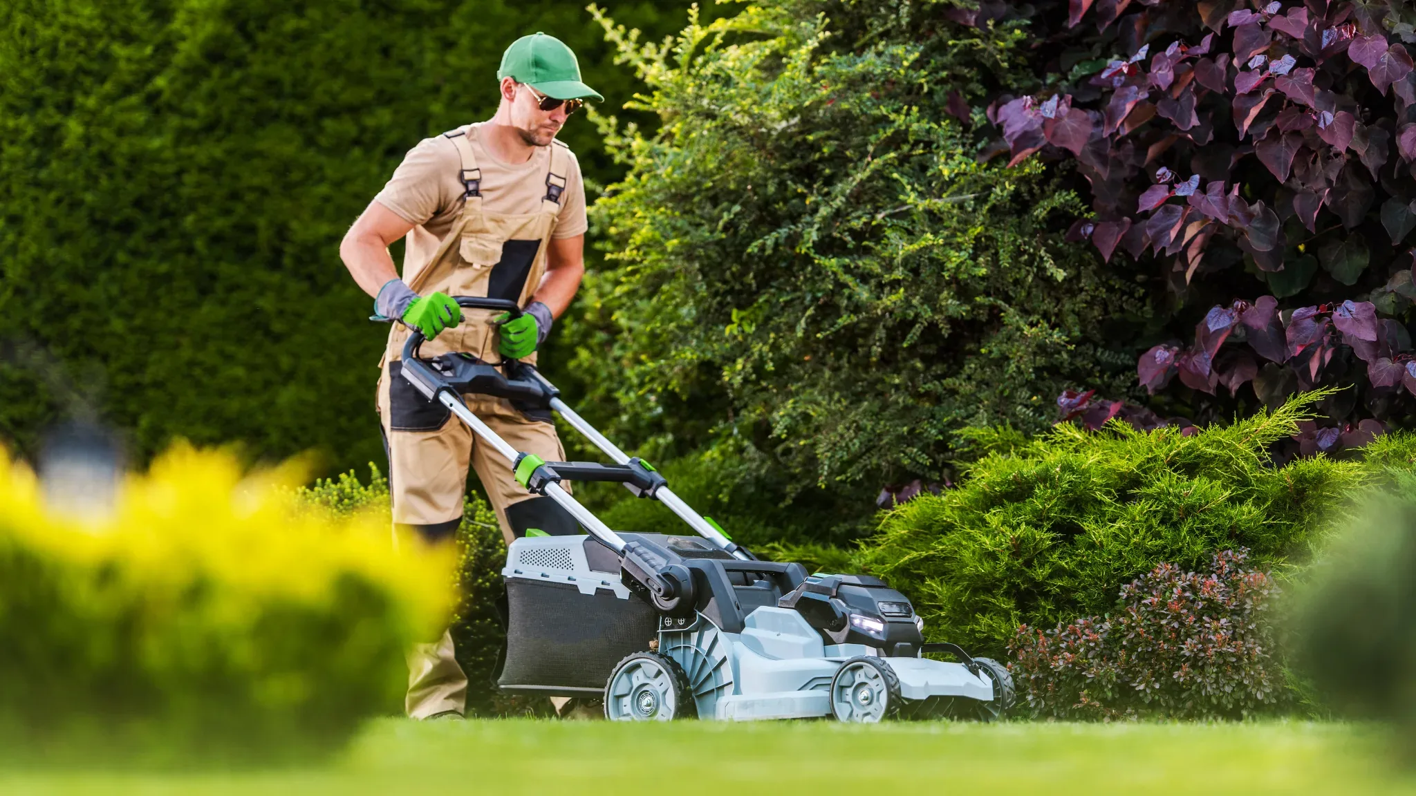 A man wearing a green cap, sunglasses, tan overalls, and green work gloves pushes a gray and black lawn mower across a green