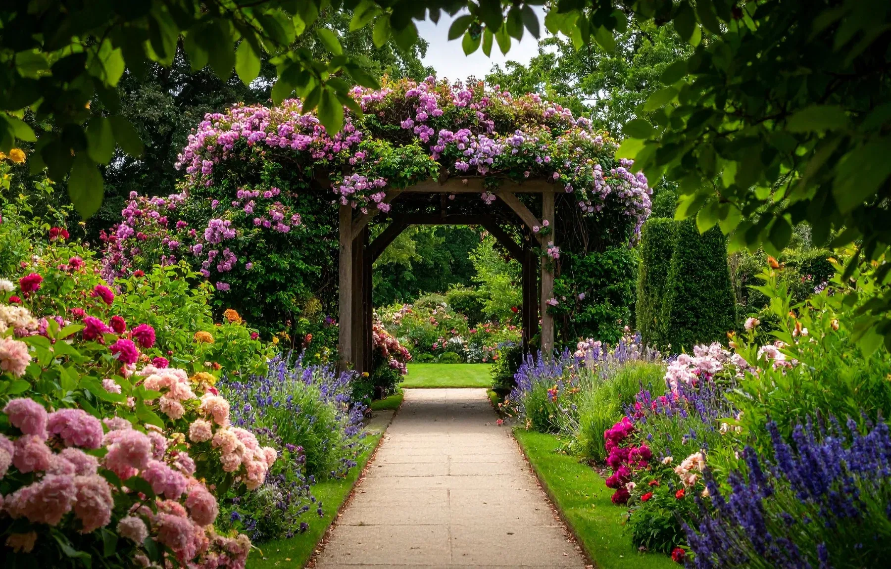 A wooden garden arbor covered in pink climbing roses arches over a paved stone path, framed by overhanging tree branches at t